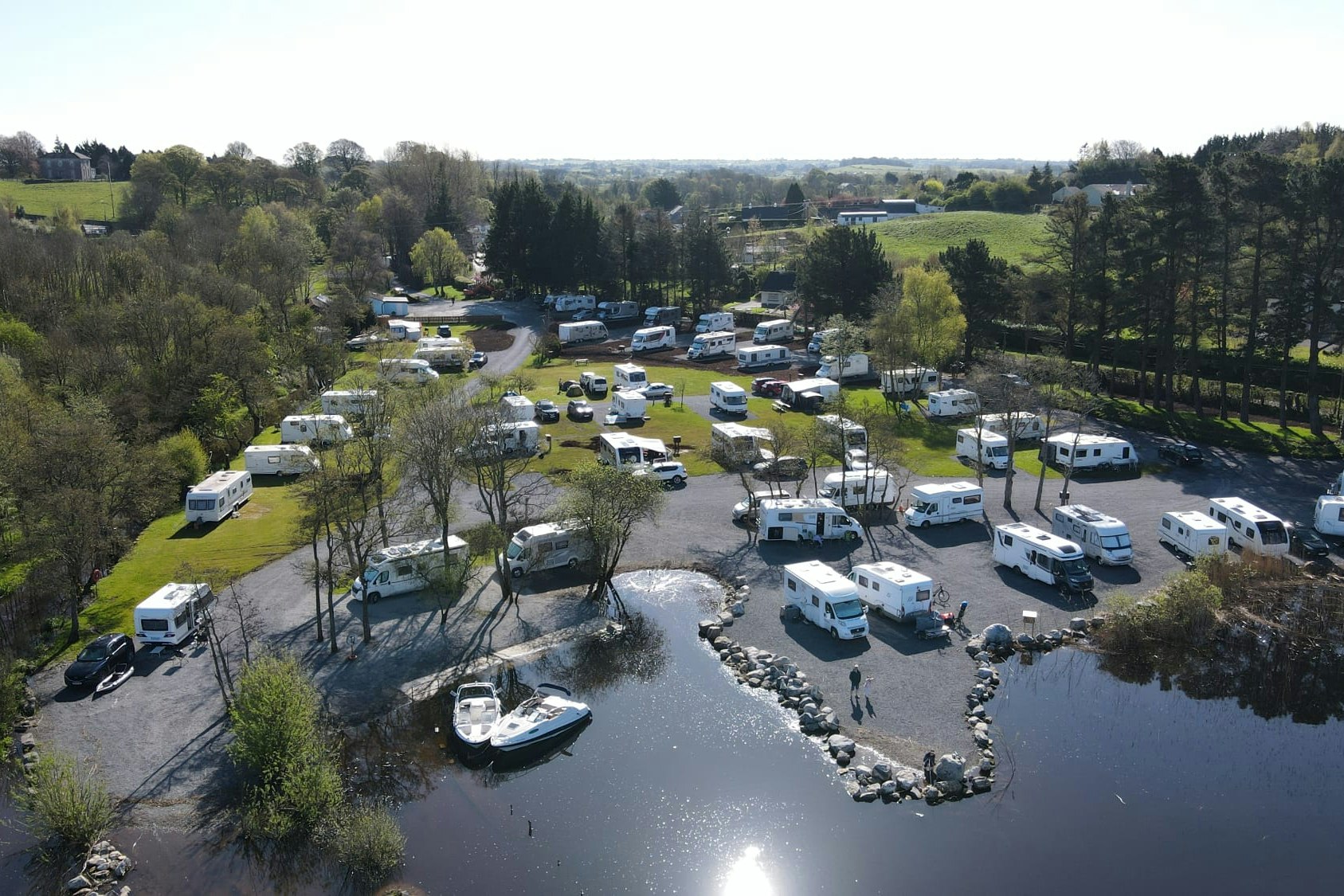 Campingplatz mit Wohnmobilen und Stellplätzen am Fluss, umgeben von Bäumen, Wegen und einem kleinen Uferbereich.