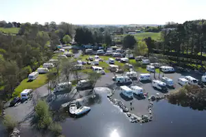 Campingplatz mit Wohnmobilen und Stellplätzen am Fluss, umgeben von Bäumen, Wegen und einem kleinen Uferbereich.