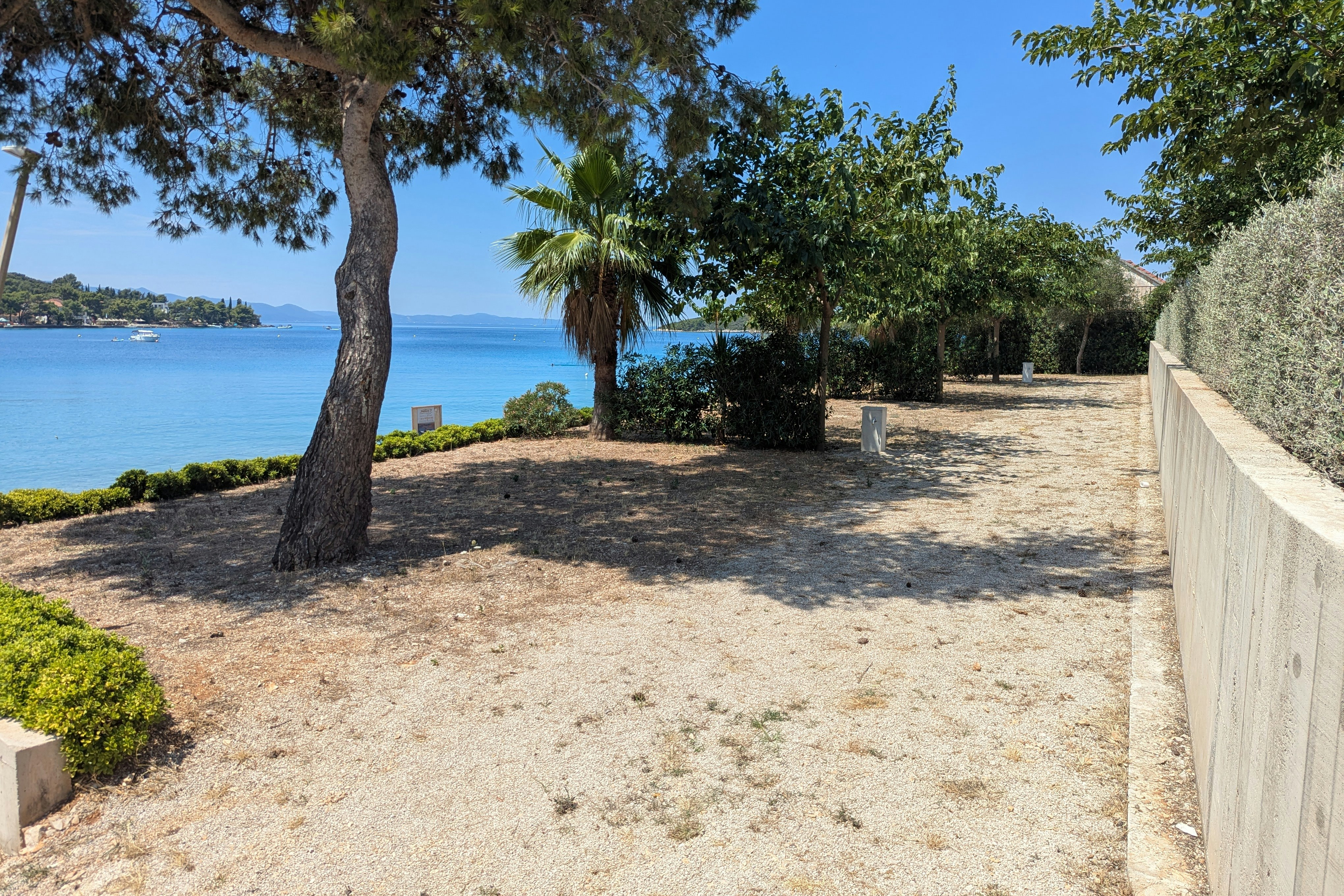 Sandiger Stellplatz am Strand mit Palmen, Bäumen und einem Holzgeländer, Blick auf das Meer und die Küste.