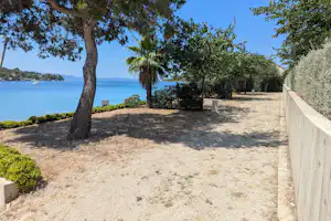 Sandiger Stellplatz am Strand mit Palmen, Bäumen und einem Holzgeländer, Blick auf das Meer und die Küste.