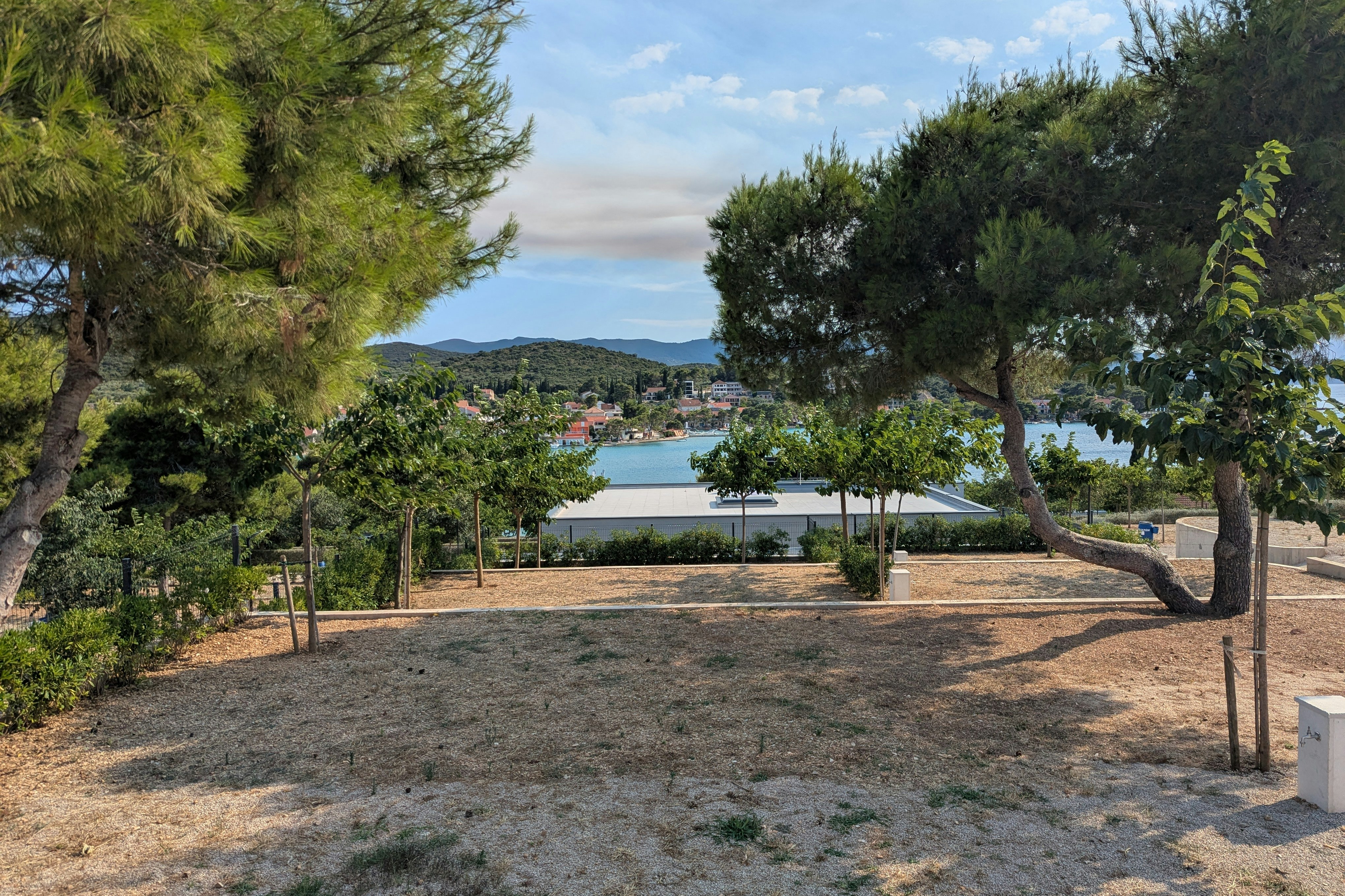 Stellplatz mit Rasenfläche und Blick auf einen See, umgeben von Bäumen und einem weißen Gebäude im Hintergrund.