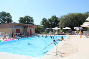 Großes Camping-Schwimmbad mit Treppe, Sonnenschirmen und Liegen, im Hintergrund Gebäude und Bäume.