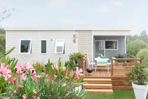 Modernes Mobilheim mit Holzdeck und Terrasse, Tisch und Stühlen, große Fensterfront und Blumenbeete im Vordergrund.