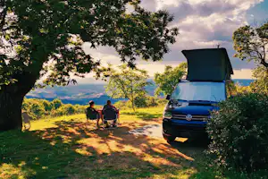 Camping-Van mit geöffneter Hecktür am Stellplatz, daneben zwei Campingstühle und Tisch unter Bäumen, Blick ins Tal.