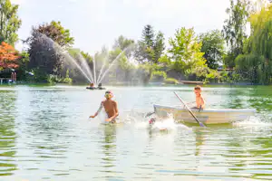 Kinder im Tretboot auf dem See, Spritzwasser und Fontänen vor Bäumen und Liegeflächen am Campingplatz.
