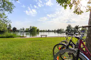 Fahrrad am Seeufer im Campingpark, Blick auf Rasen, Bank, Springbrunnen und Bäume am Wasser.
