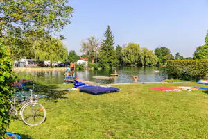 Grüne Wiese am Seeufer mit Liegen, bunten Schwimmdecken, Fahrrad und Personen im Hintergrund.