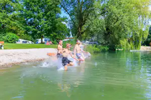 Badende Personen im See vor dem Campingplatz, im Hintergrund Wohnwagen und Zelte am Ufer unter Bäumen.