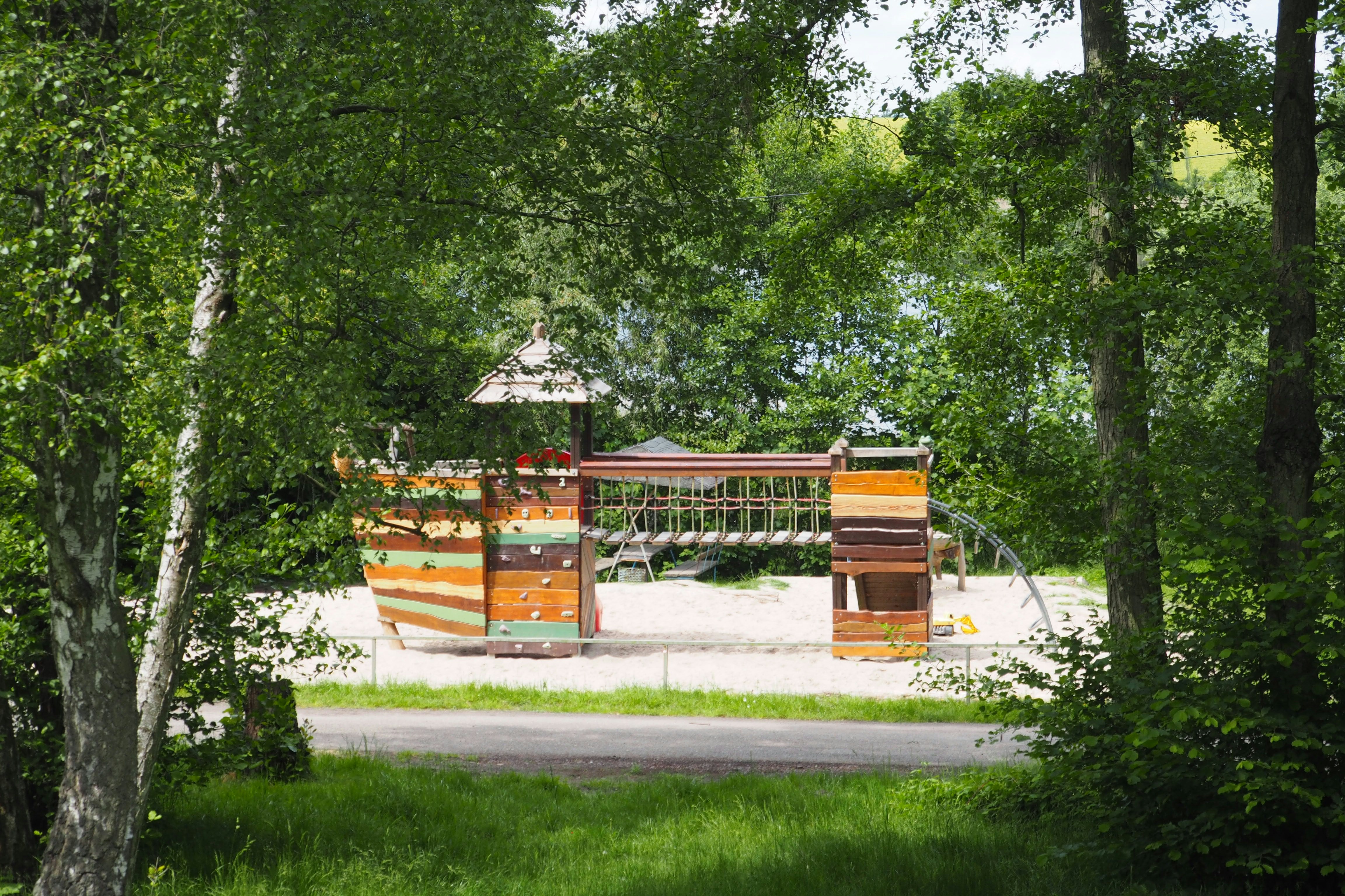 Buntes Spielhaus und Sitzbereich auf einem Campingplatz, umgeben von Bäumen und Rasen, mit Sandfläche im Hintergrund.