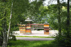 Buntes Spielhaus und Sitzbereich auf einem Campingplatz, umgeben von Bäumen und Rasen, mit Sandfläche im Hintergrund.