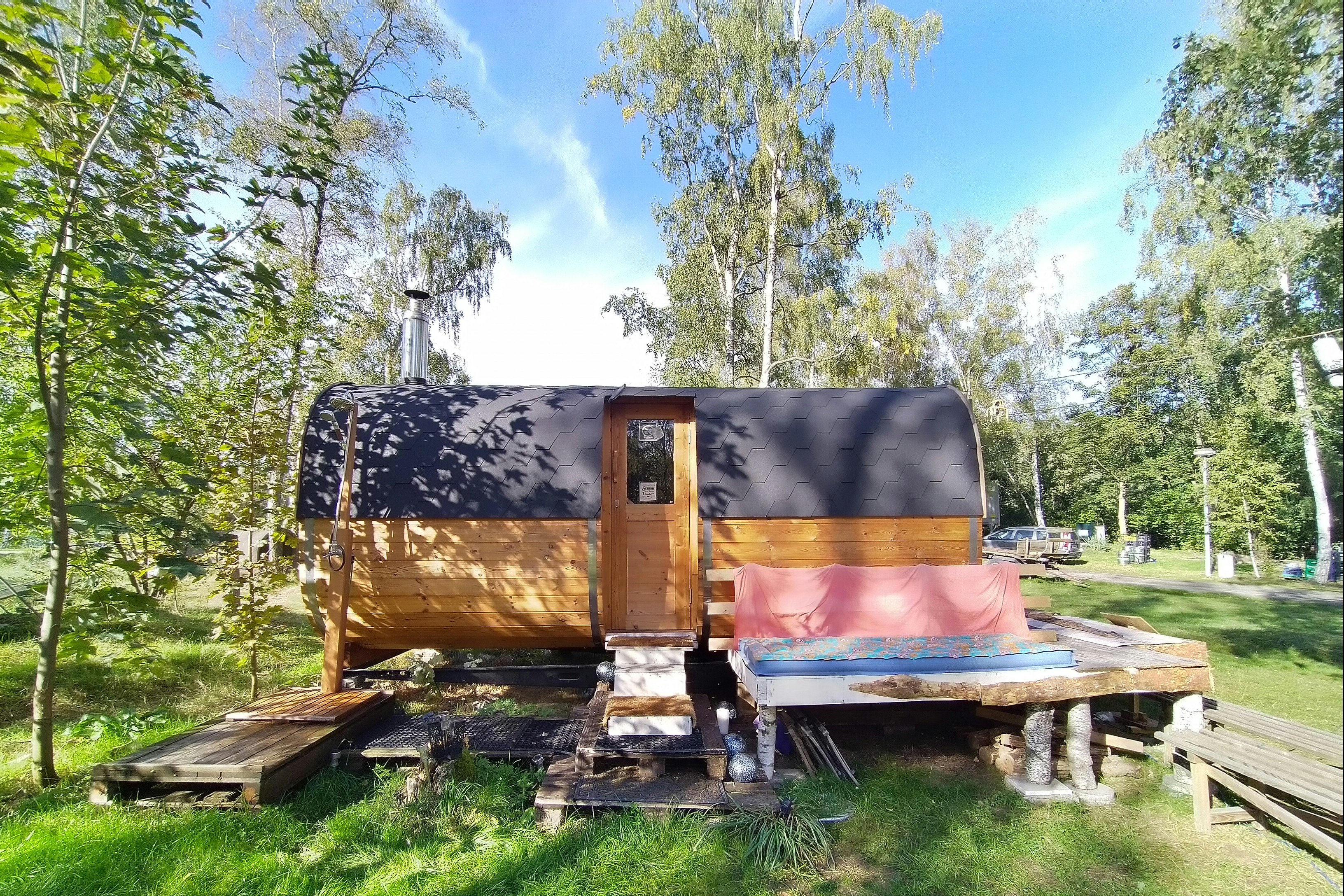 Holzanhänger mit Plane und geöffnetem Aufbau auf einem Campingplatz, umgeben von Gras, Bäumen und einem Holzpodest.