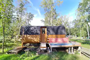 Holzanhänger mit Plane und geöffnetem Aufbau auf einem Campingplatz, umgeben von Gras, Bäumen und einem Holzpodest.