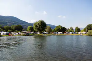 Seeufer mit Wohnwagen und Campingplätzen am Rand, dahinter Bäume und Hügel in der Ferne.