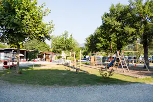 Spielplatz auf dem Campingplatz mit Schaukeln, Holzgerüst und Rasenfläche, umgeben von Bäumen und Wegen.