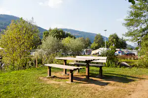 Holztisch und Sitzbank auf einer Wiese im Campingbereich, im Hintergrund Olivenbäume, Wohnwagen und Berge.