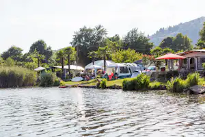 Campingplatz am See mit Wohnwagen, Zelt und Sonnenschirmen am Ufer, umgeben von Büschen und Bäumen.