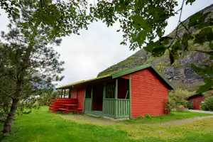Rote Holzhütte mit grünen Türen und Veranda am Campingplatz, Grasweg und Bäume vor einer steilen Felswand.