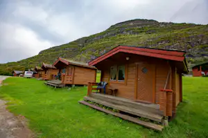 Holz-Chalet mit Veranda und Stufen auf einer grünen Wiese, weitere Chalets am Weg, im Hintergrund ein bewachsener Hügel.
