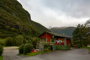 Rotes Campinghaus mit Veranda, Sitzbank und Weg am Rand, umgeben von Bäumen und grünen Bergen im Hintergrund.