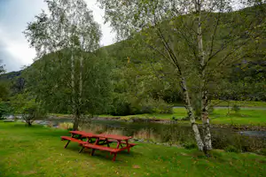 Grüner Campingplatz mit roter Picknickbank und Bäumen am Bach, im Hintergrund Wiesen und bewaldete Hügel.