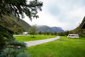 Wohnwagen auf einem Campingplatz auf einer grünen Wiese, mit Kiesweg, Bäumen und Bergen im Hintergrund.