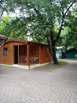 Holzhaus auf einem Campingplatz mit Veranda, Zäunen und gepflastertem Weg, umgeben von großen Bäumen im Grünen.