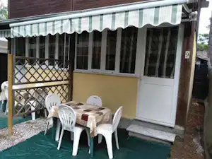 Wohnwagen mit Terrasse, Tisch und weißen Stühlen unter einem gestreiften Markisen-Vordach, daneben Holzgeländer und Eingangstür.