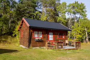 Holzhütte im Campingbereich mit Terrasse, Sitzbank und Tisch vor Waldkulisse.