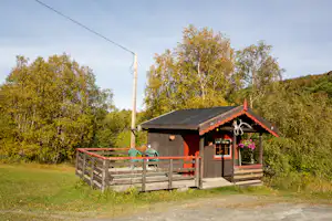 Holzhaus mit Terrasse und rotem Geländer, umzäunter Fläche und Wiese im Vordergrund, umgeben von Bäumen im Hintergrund.