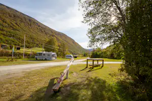 Wohnmobil auf Campingplatz am Weg, Wiese mit Bank und Absperrpfosten, Berge im Hintergrund und Bäume am Rand.