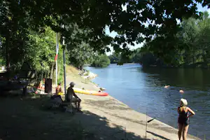 Camping am Flussufer unter Bäumen: Kajak am Steg, Personen am Rand und ein Boot auf dem Wasser.