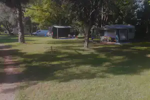 Campingplatz mit Wohnwagen und Zelt unter Bäumen, davor Campingstühle und Tisch auf dem Gras.