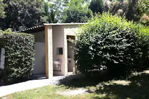 Camping-Bungalow mit Terrasse, Waschbecken und Dusche im offenen Bereich, umgeben von Hecken und Bäumen.