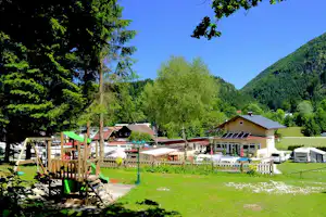 Campingplatz mit grüner Wiese, Ferienhäusern, Solardach, Wohnwagen und Wohnmobilen, umgeben von Bäumen und Hügeln.
