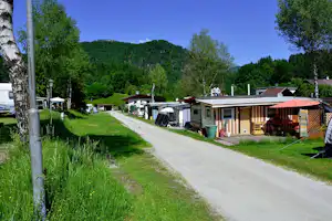 Campingplatz mit Wohnwagen und Mobilheimen entlang eines breiten Wegs, grüner Wiese, Bäumen und Bergen im Hintergrund.
