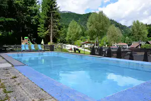 Schwimmbecken auf dem Campingplatz mit blauem Wasser, Liegefläche und Blick auf Wohnwagen, Zaun und Bäume im Grünen.