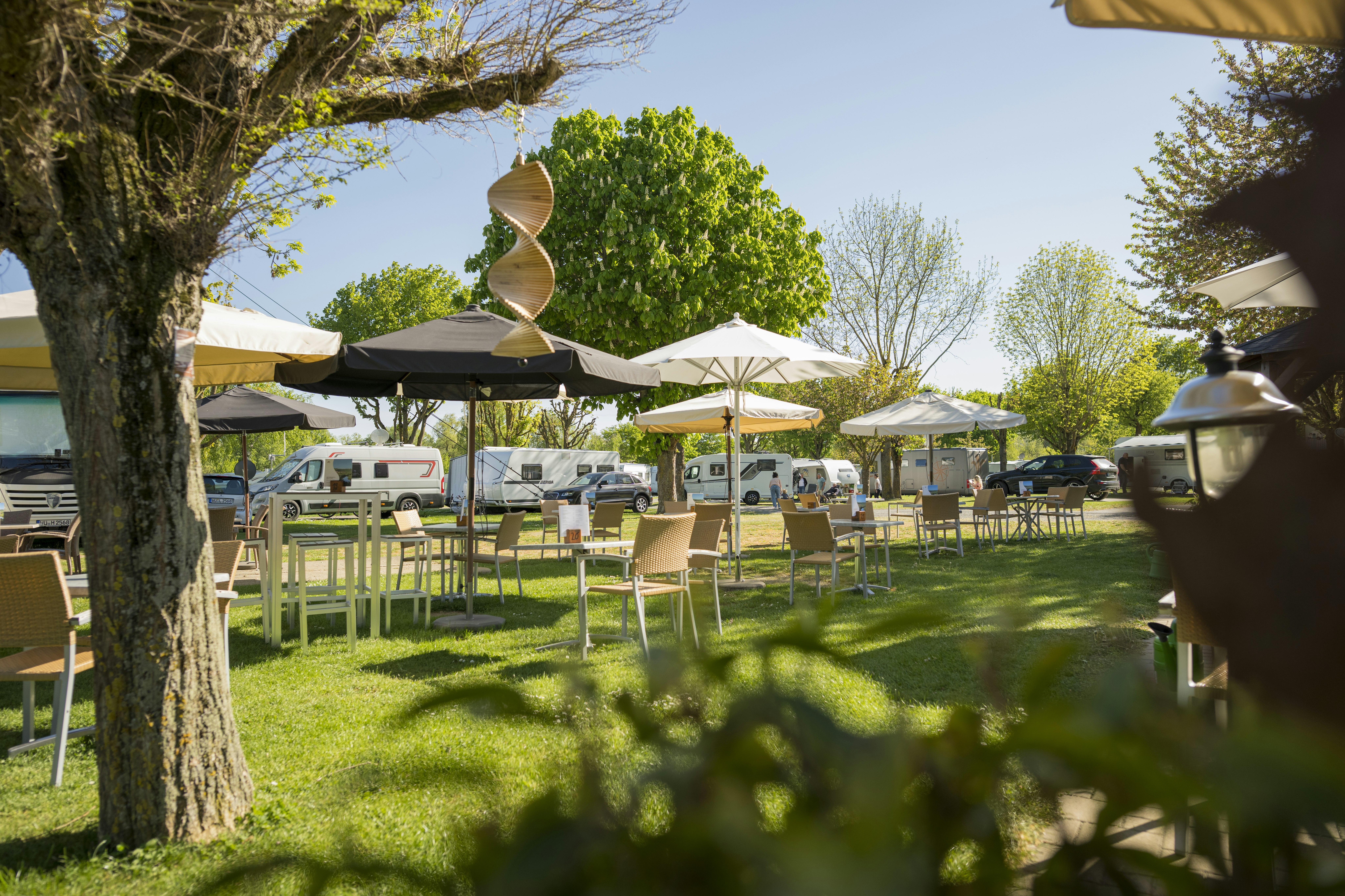 Grüner Campingplatz mit Rasen, Holztischen und Stühlen unter Sonnenschirmen, im Hintergrund Wohnmobile und Zelte zwischen Bäumen.