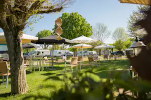 Grüner Campingplatz mit Rasen, Holztischen und Stühlen unter Sonnenschirmen, im Hintergrund Wohnmobile und Zelte zwischen Bäumen.
