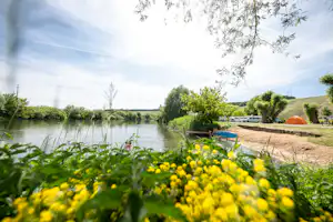 Blick auf den See am Campingplatz, im Vordergrund Blumenbeet, daneben Rasen und ein orangefarbenes Zelt unter Bäumen.