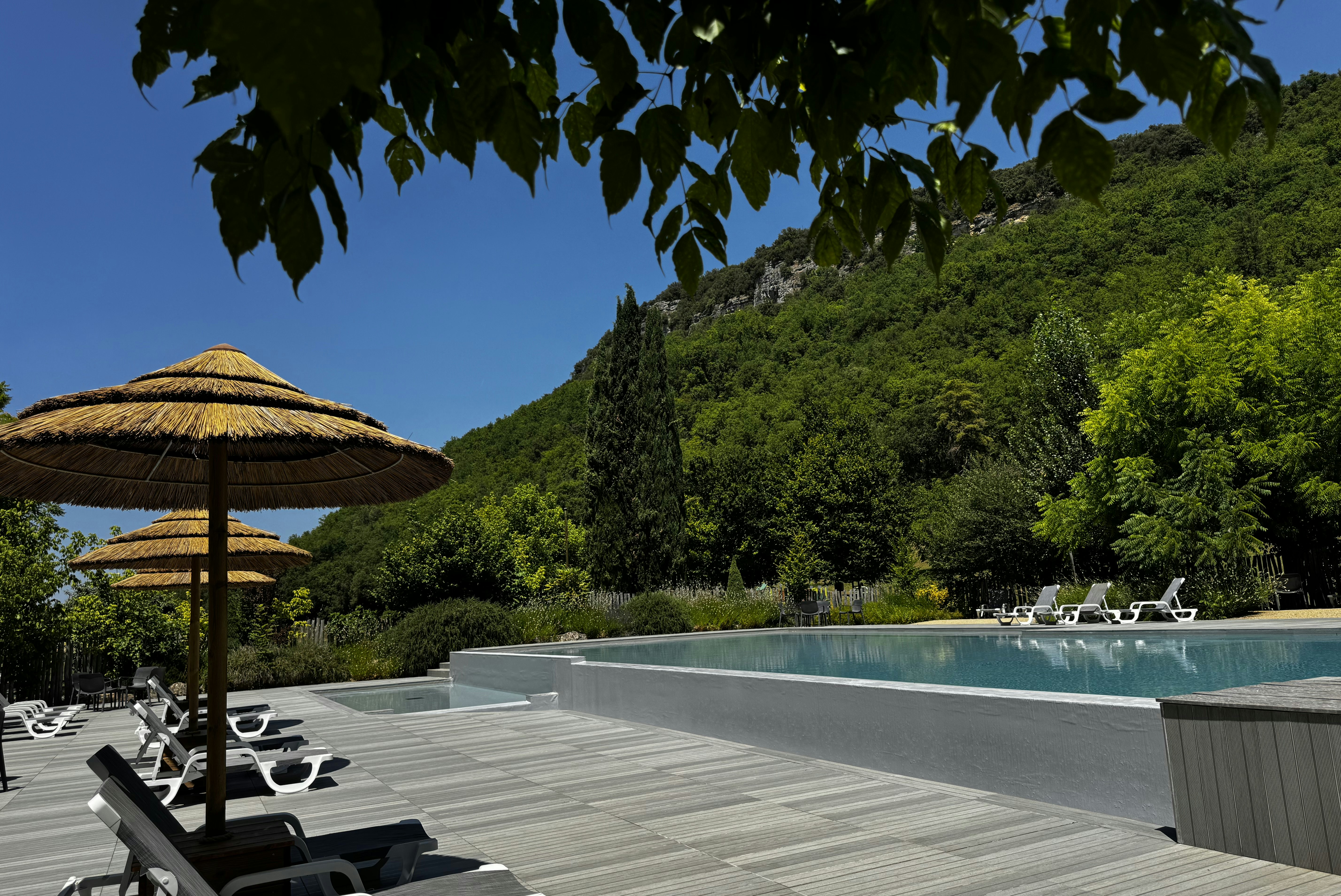Camping-Pool mit Liegestühlen auf Holzterrasse, daneben Sonnenschirme und dichtes Grün vor blauem Himmel.
