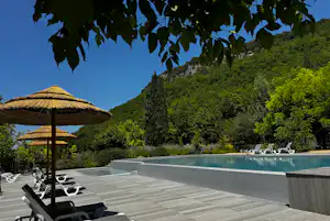 Camping-Pool mit Liegestühlen auf Holzterrasse, daneben Sonnenschirme und dichtes Grün vor blauem Himmel.