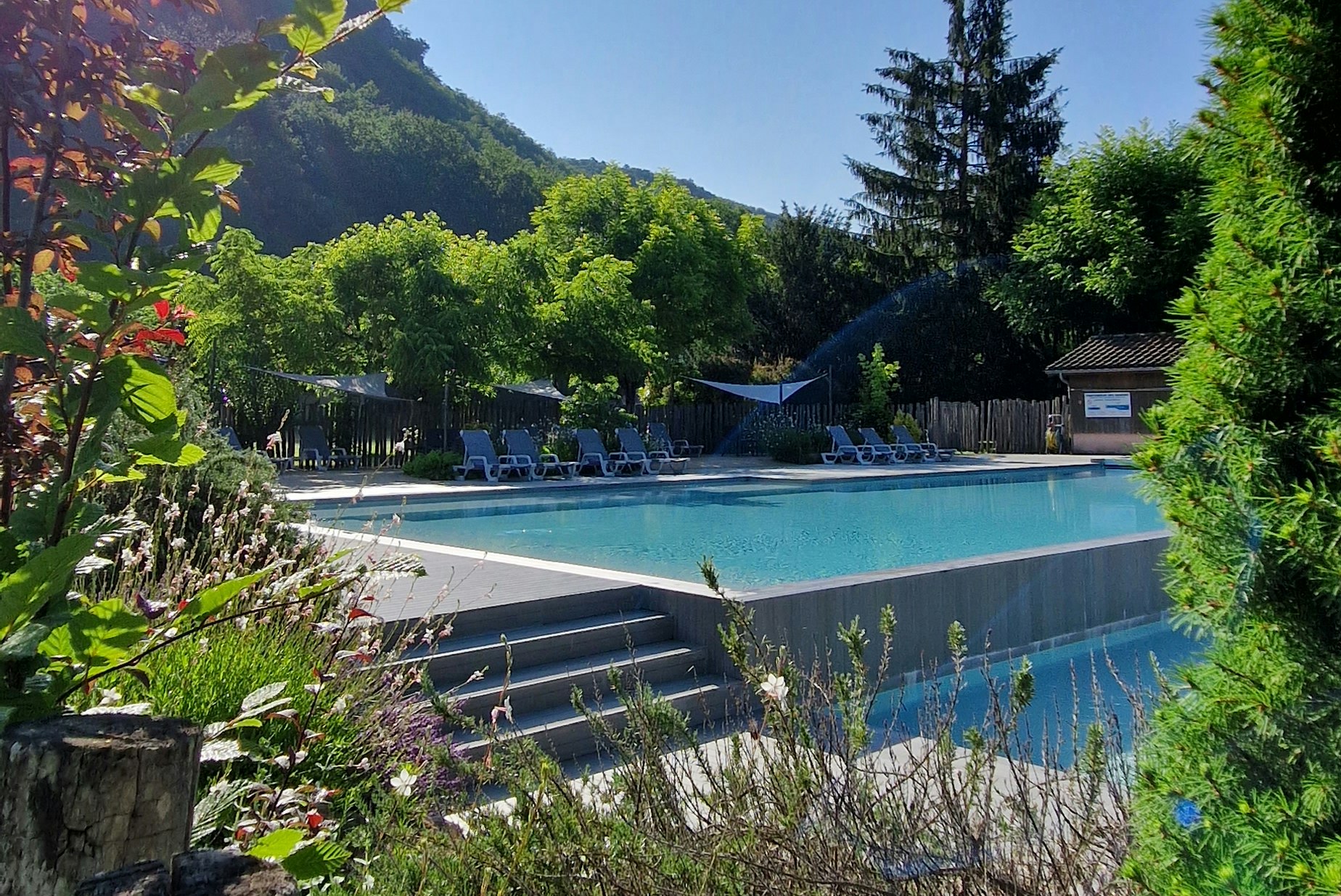 Schwimmbecken im Campingbereich mit Liegestühlen, umgeben von Grün und Bergen im Hintergrund.