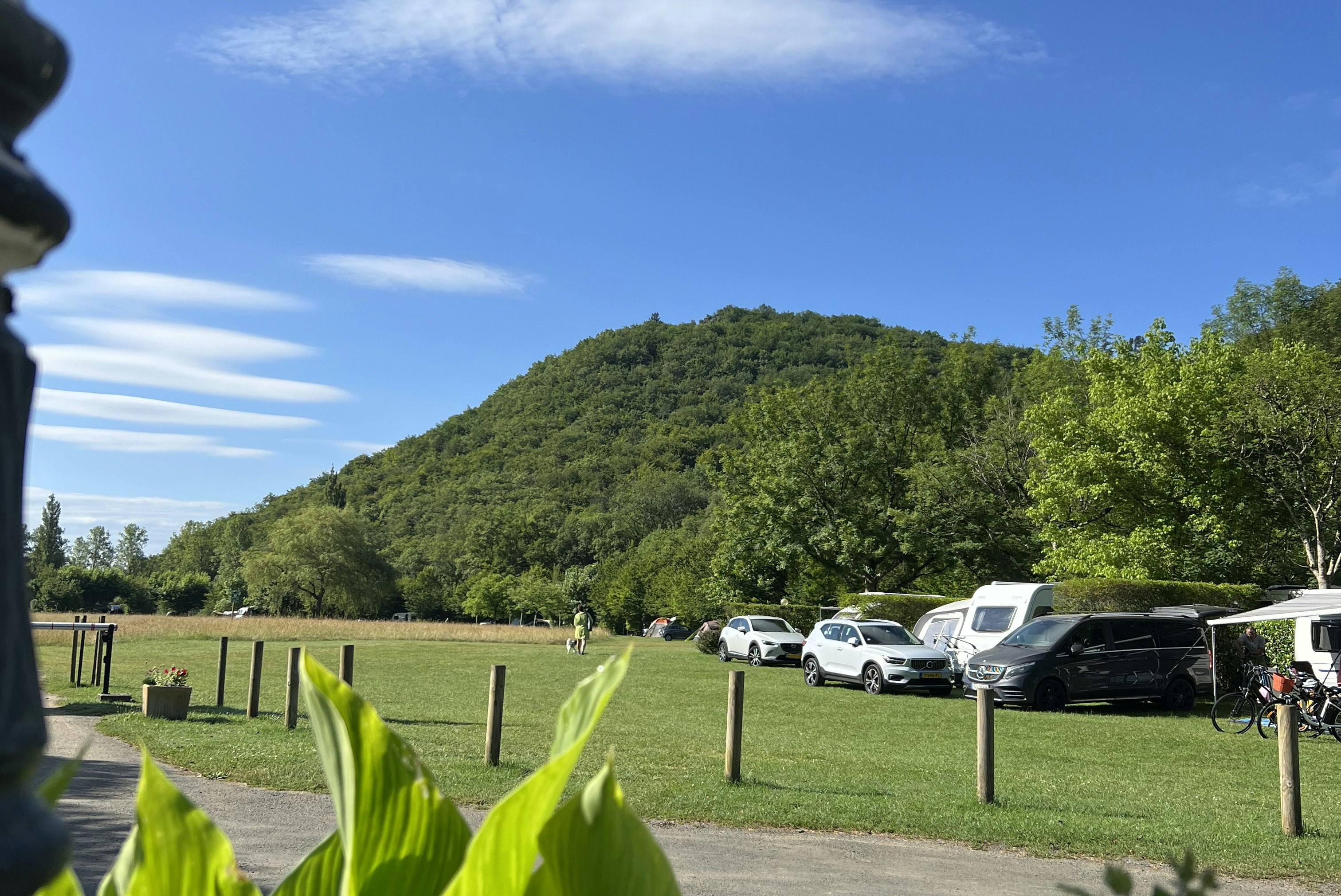 Campingplatz mit grünem Rasen, geparkten Autos und Wohnwagen, im Hintergrund ein bewaldeter Hügel und blauem Himmel.