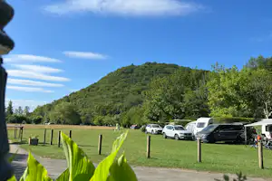 Campingplatz mit grünem Rasen, geparkten Autos und Wohnwagen, im Hintergrund ein bewaldeter Hügel und blauem Himmel.
