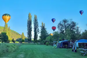 Campingwiese mit mehreren Zelten am Rand, Bänke und hohe Bäume, im Hintergrund fliegen Heißluftballons am blauen Himmel.
