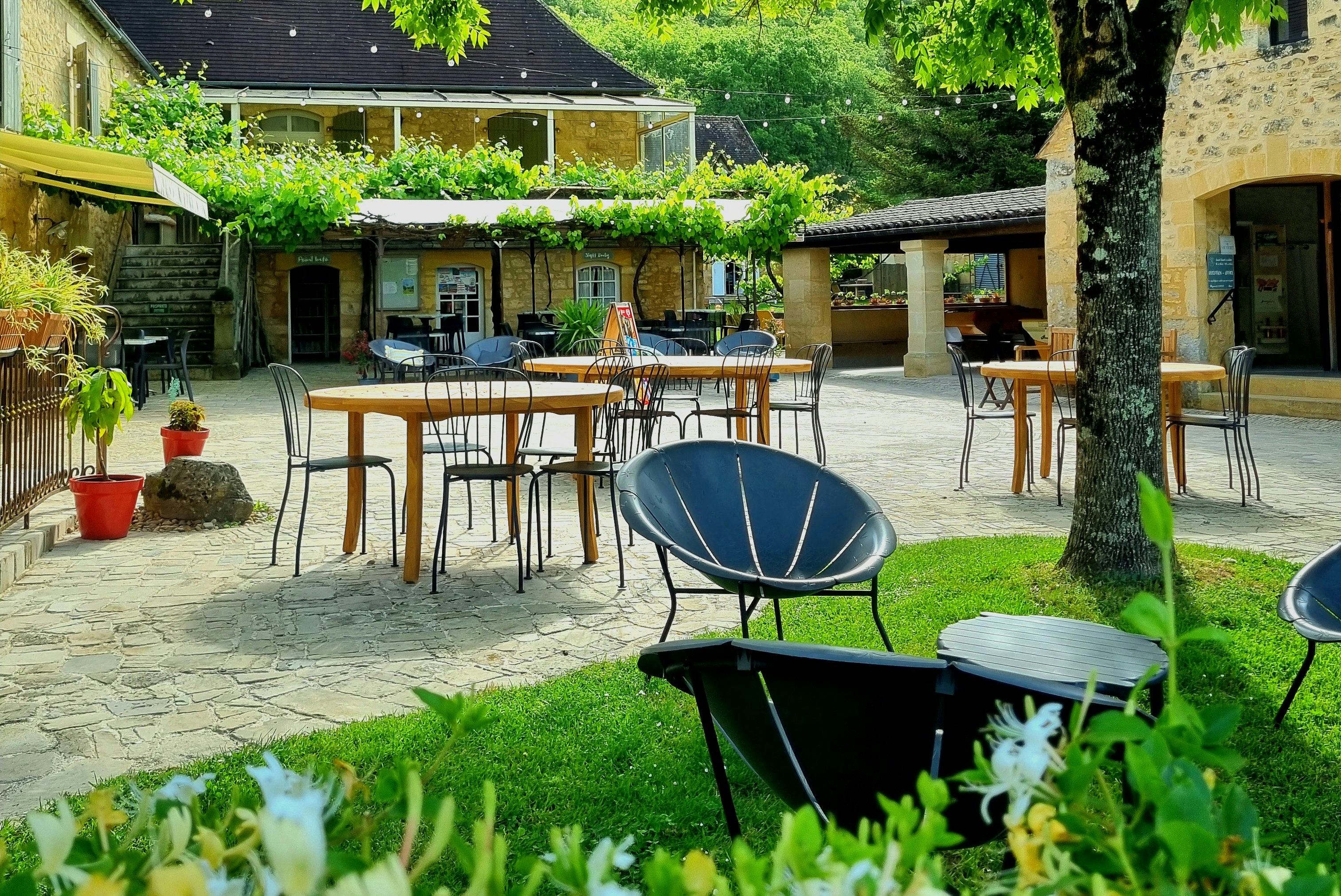 Terrasse im Campinghof mit Tischen und Stühlen, Grill und Sonnenschirm, umgeben von Bäumen und Grünpflanzen.