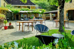Terrasse im Campinghof mit Tischen und Stühlen, Grill und Sonnenschirm, umgeben von Bäumen und Grünpflanzen.