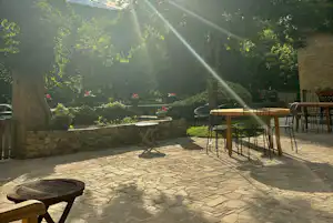 Camping-Terrasse mit Tischen und Stühlen, Steinmauer und Bäumen im Hintergrund, Sonnenstrahlen am Himmel.