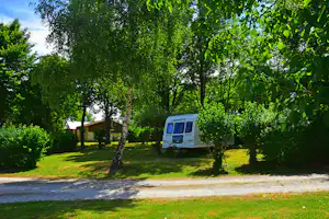 Wohnwagen auf einem grünen Campingplatz zwischen Bäumen, mit gepflastertem Weg und Rasenfläche im Hintergrund.