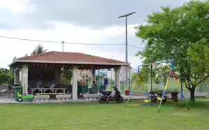 Spielplatz mit Schaukel und Rasen vor einem Campinghaus mit überdachter Terrasse, Tischen, Stühlen und Motorroller.